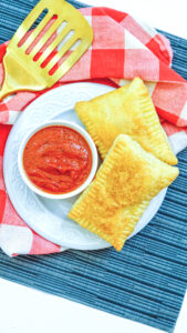 A blue placemat topped with a red and white checked cloth napkin with a plate of two calzones and a small bowl of pizza sauce.