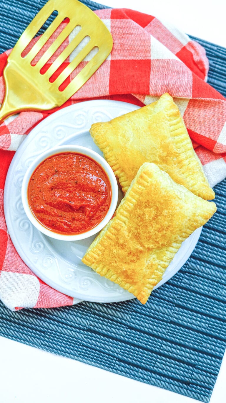 A blue placemat topped with a red and white checked cloth napkin with a plate of two calzones and a small bowl of pizza sauce.