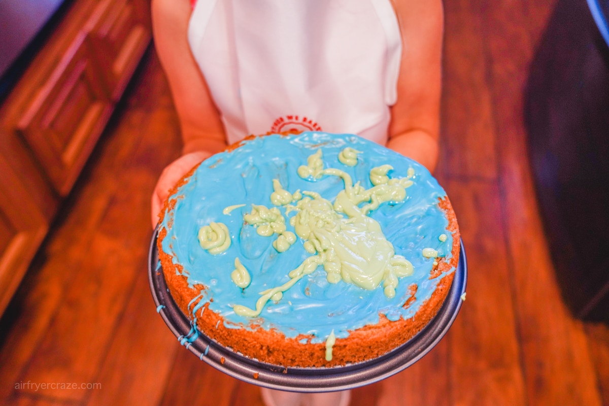A small child holding a cookie cake with blue and green icing frosted on top.