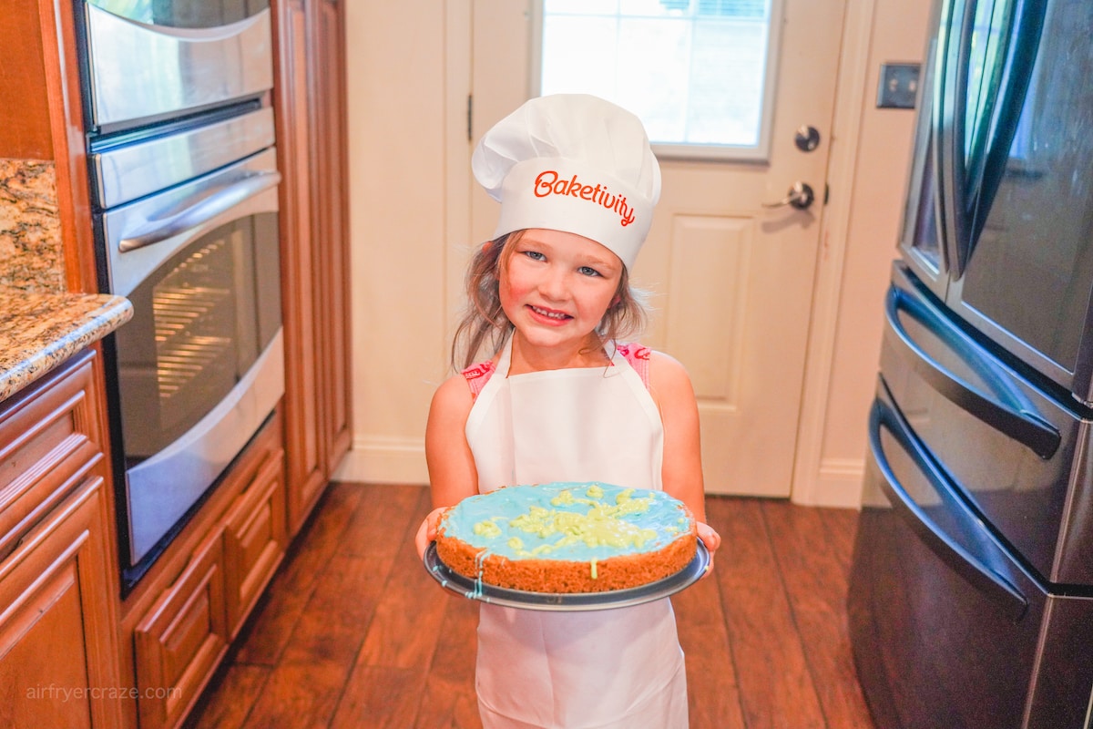 A little girl in a chef hat and apron, holding a giant chocolate chip cookie decorated to look like Earth.