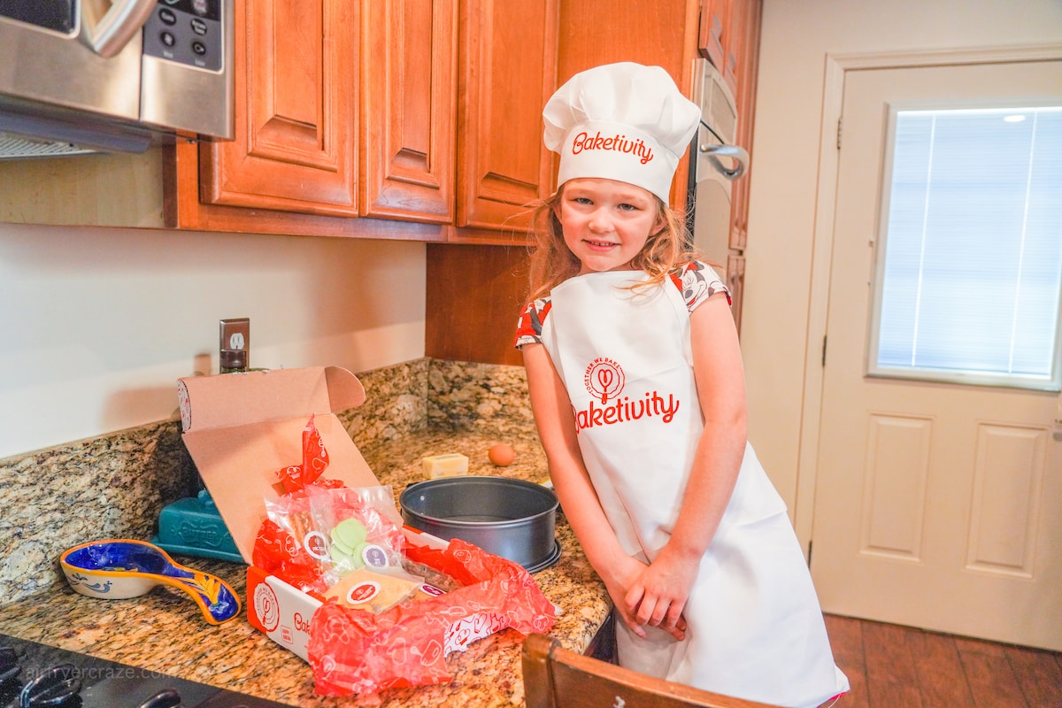 A girl in an apron and chef hat, posing next to an open Baketivity Box.