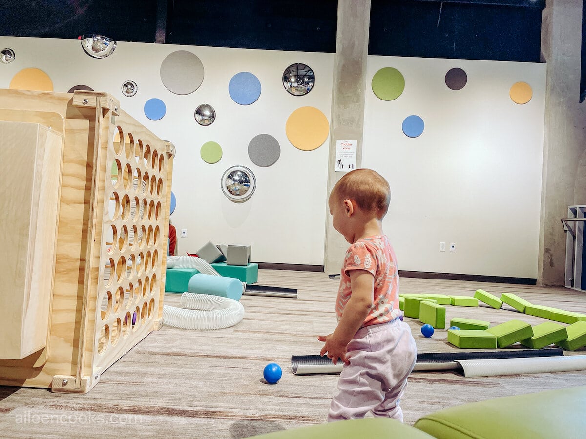 A toddler inside the toddler zone that's filled with balls and large, soft building blocks.