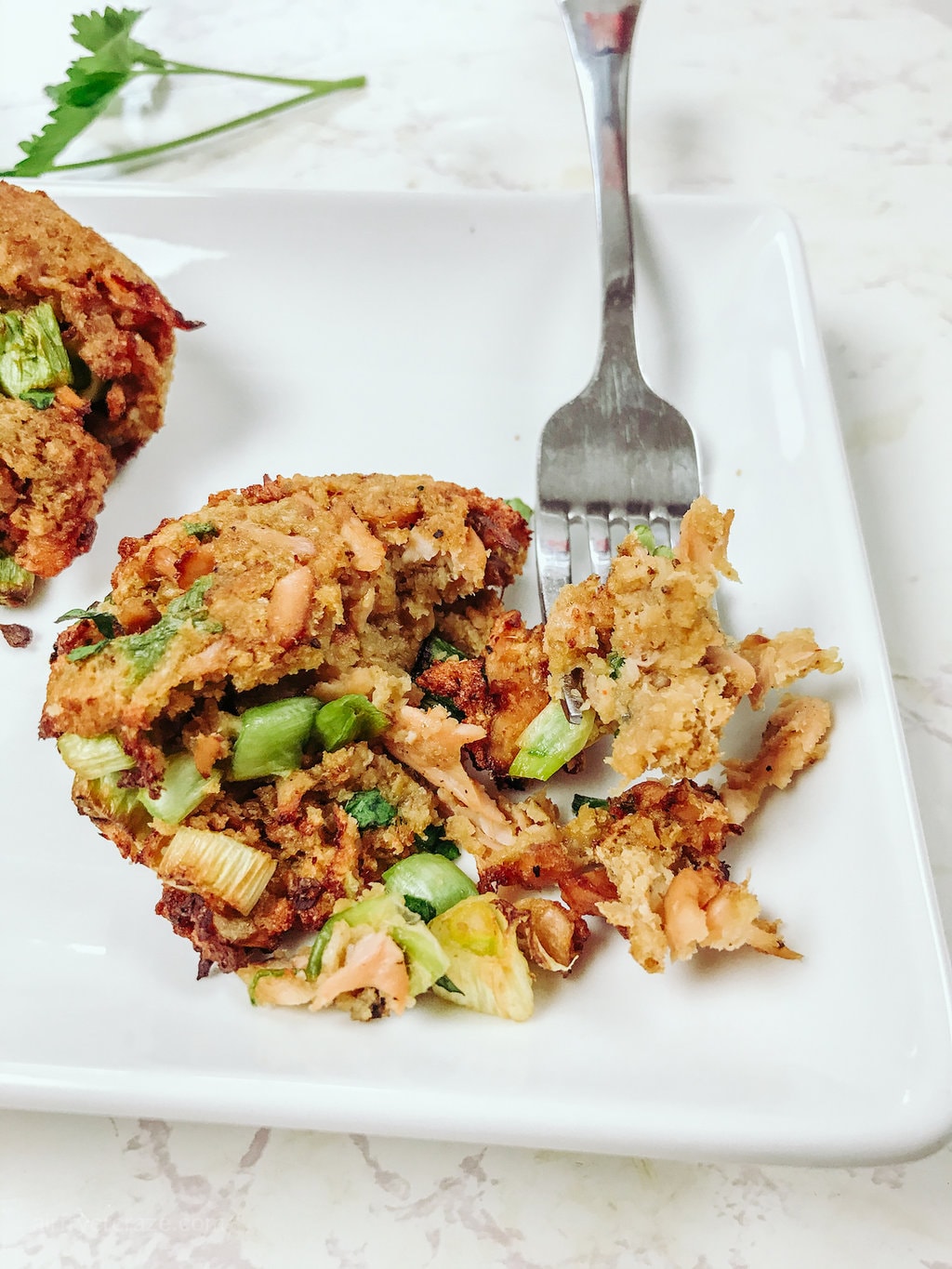 A white, rectangular plate, filled with delicious crispy salmon cakes and garnished with parsley. One salmon cake is being cut through with a fork.