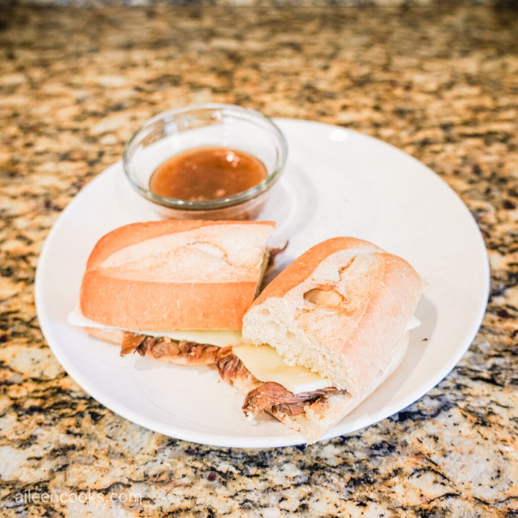 Crock Pot French Dip on a white plate, cut in half and with a small glass bowl of au jus next to it