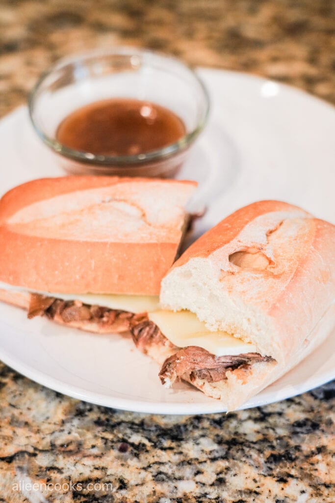 Crock Pot French Dip on a white plate, cut in half and with a small glass bowl of au jus next to it