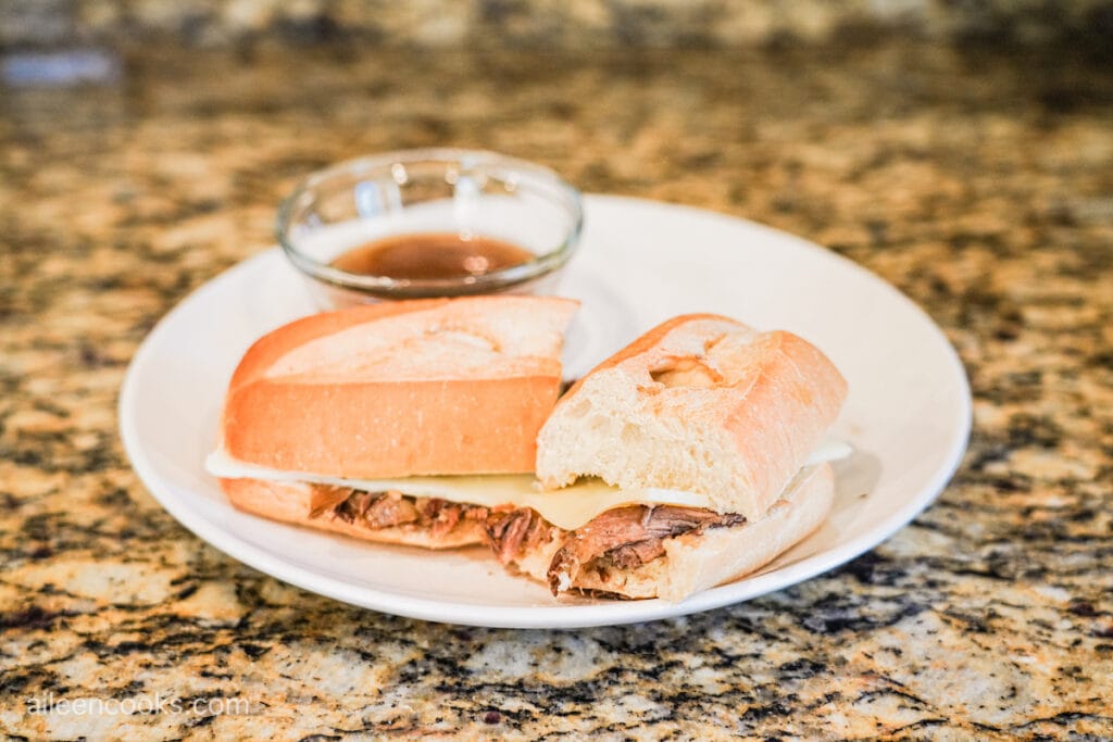 Crock Pot French Dip on a white plate, cut in half and with a small glass bowl of au jus next to it