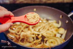 Minced garlic in a red measuring spoon, being added to a skillet of sliced onions
