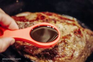 Worcestershire sauce in a red measuring spoon, being added to browned chuck roast
