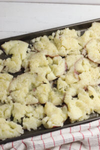 Smashed potatoes on a black sheet pan and seasoned with salt and pepper, sitting on a red and white plaid tea towel