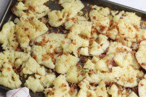 Smashed potatoes on a black sheet pan, seasoned with salt and pepper, and covered with bits of bacon