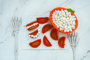 A process photo of vampire teeth cookies being made: halved sugar cookies with red frosting on a white cutting board. A large pumpkin-shaped bowl filled with mini marshmallows sit beside them.