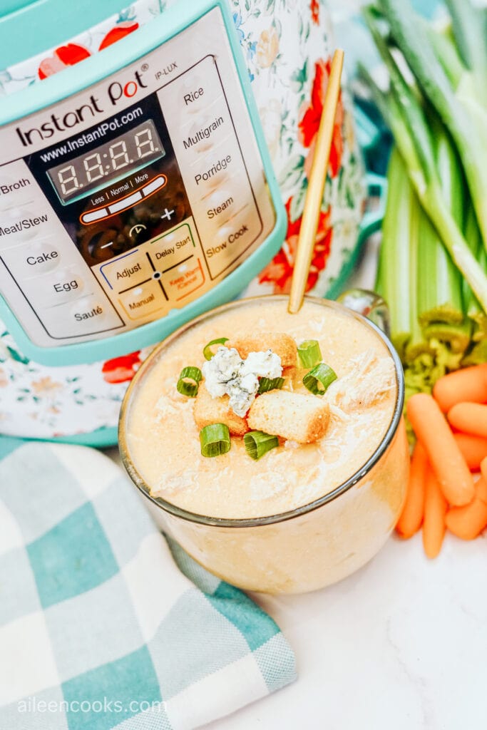 A cup of Buffalo Chicken Soup, garnished with croutons, blue cheese and green onions, sitting in front of an Instant Pot machine, surrounded by stalks of celery and carrots.