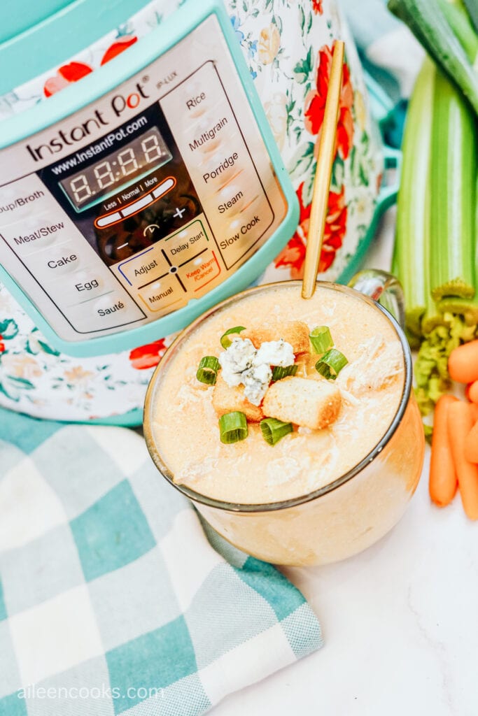 Buffalo chicken soup in a glass cup, fully garnished, sitting in front of a teal-colored Instant Pot.