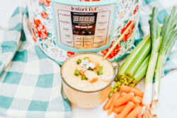 A glass cup of chicken soup, in front of a floral-wrapped Instant Pot, surrounded by a blue plaid tea towel, stalks of celery and baby carrots.