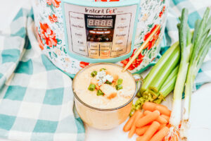 A glass cup of chicken soup, in front of a floral-wrapped Instant Pot, surrounded by a blue plaid tea towel, stalks of celery and baby carrots.