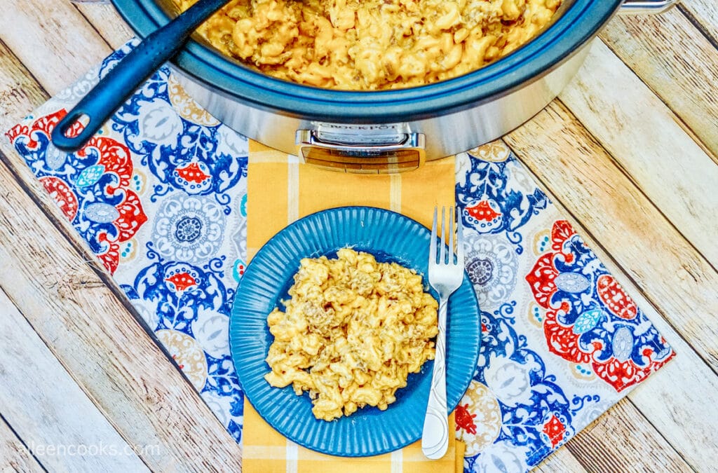 Bird’s eye view of a serving of cheeseburger mac and cheese served on a blue plate with a fork, sitting on a beautiful table laid out with a yellow table runner and blue patterned place mat. A Crockpot full of more mac and cheese sits in the background.