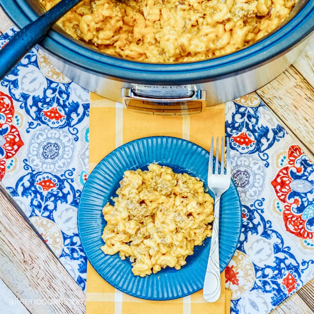 Aerial view of a serving of cheeseburger mac and cheese served on a blue plate with a fork, sitting on a beautiful table laid out with a yellow table runner and blue patterned place mat. A Crockpot full of more mac and cheese sits in the background.