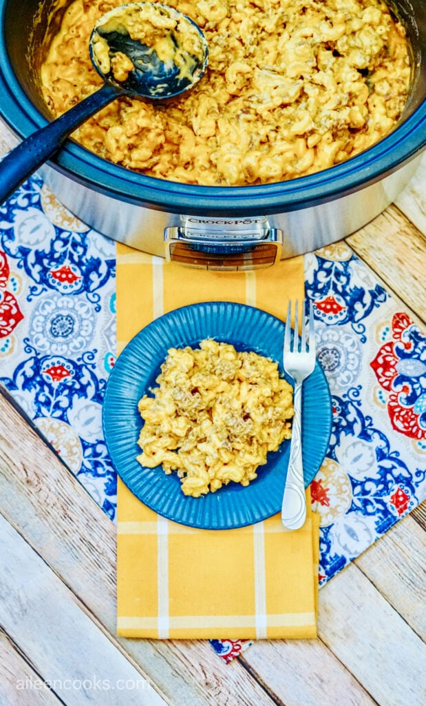 Bird’s eye view of a serving of cheeseburger mac and cheese served on a blue plate with a fork, sitting on a beautiful table laid out with a yellow table runner and blue patterned place mat. A Crockpot full of more mac and cheese sits in the background.