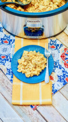 A serving of cheeseburger mac and cheese on a blue plate, a fork, and a slow cooker filled with more macaroni