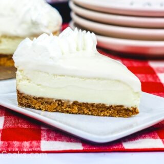 A slice of Instant Pot Cheesecake sitting on a square white plate, sitting on a plaid red tablecloth. A stack of plates and the whole cheesecake sit in the background.