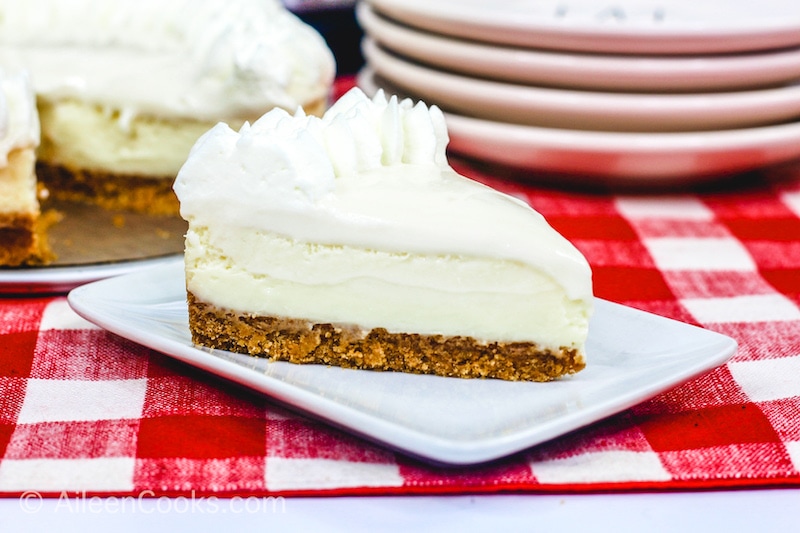 A slice of Instant Pot Cheesecake sitting on a square white plate, sitting on a plaid red tablecloth. A stack of plates and the whole cheesecake sit in the background.