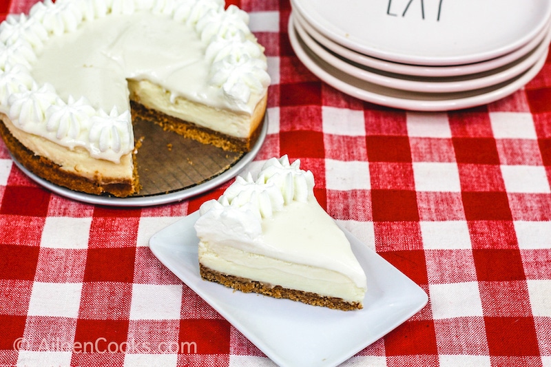 A bird’s eye view of a slice of Instant Pot Cheesecake sitting on a square white plate, sitting on a plaid red tablecloth. A stack of plates and the whole cheesecake sit in the background.