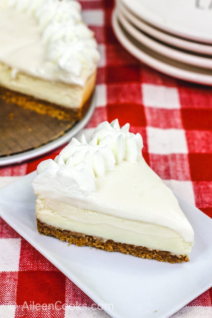 A bird’s eye view of a slice of Instant Pot Cheesecake sitting on a square white plate, sitting on a plaid red tablecloth. A stack of plates and the whole cheesecake sit in the background.