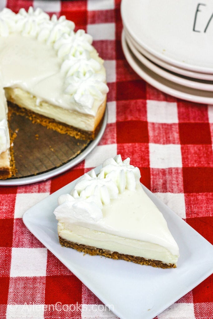 A bird’s eye view of a slice of Instant Pot Cheesecake sitting on a square white plate, sitting on a plaid red tablecloth. A stack of plates and the whole cheesecake sit in the background.