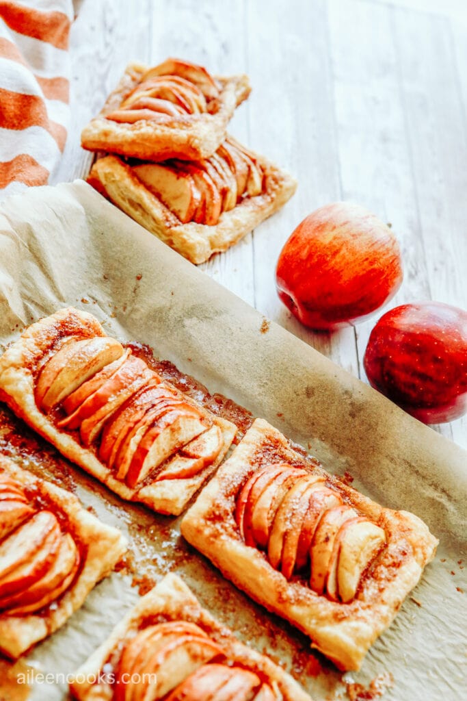 Mini apple tarts sitting on a parchment-lined baking sheet, surrounded by two additional tarts and two red apples.