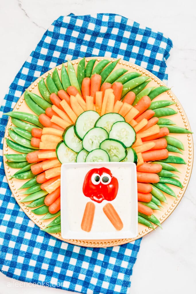 Cucumbers, orange bell peppers, baby carrots and snap peas laid out on a large circle tray to make up a Turkey Veggie Tray, with a blue plaid tea towel in the background.
