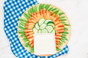 Vegetables including cucumber and snap peas, laid out in a circular form, with a square-shaped bowl filled with ranch sauce.