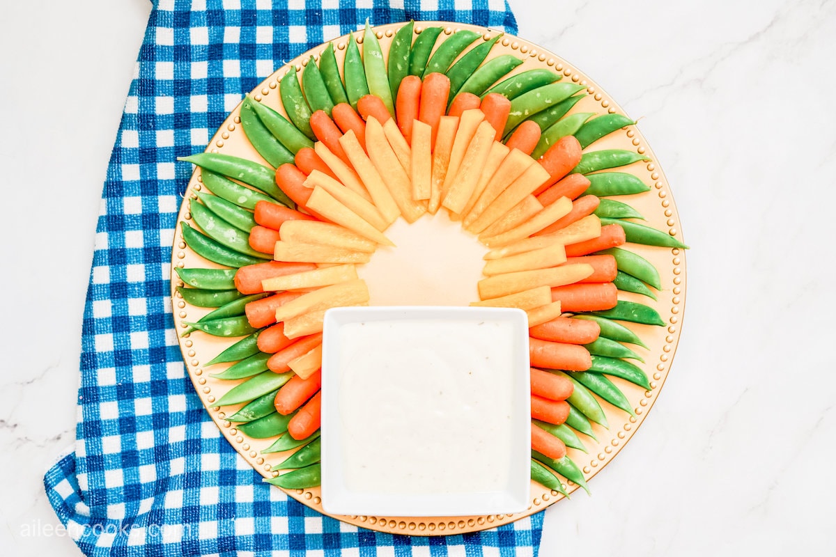 Orange bell pepper, baby carrots, and snap peas layered on a circular tray with a square-shaped bowl of ranch sauce
