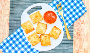 Air fryer ravioli on a white plate served with marinara sauce and a gold fork, surrounded by bowls of additional ingredients, sitting on a blue plaid napkin
