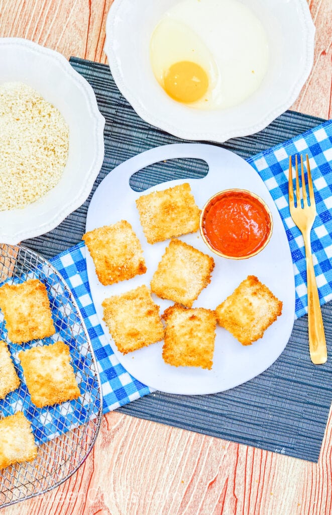 Air fryer ravioli on a white plate served with marinara sauce and a gold fork, surrounded by bowls of additional ingredients, sitting on a blue plaid napkin