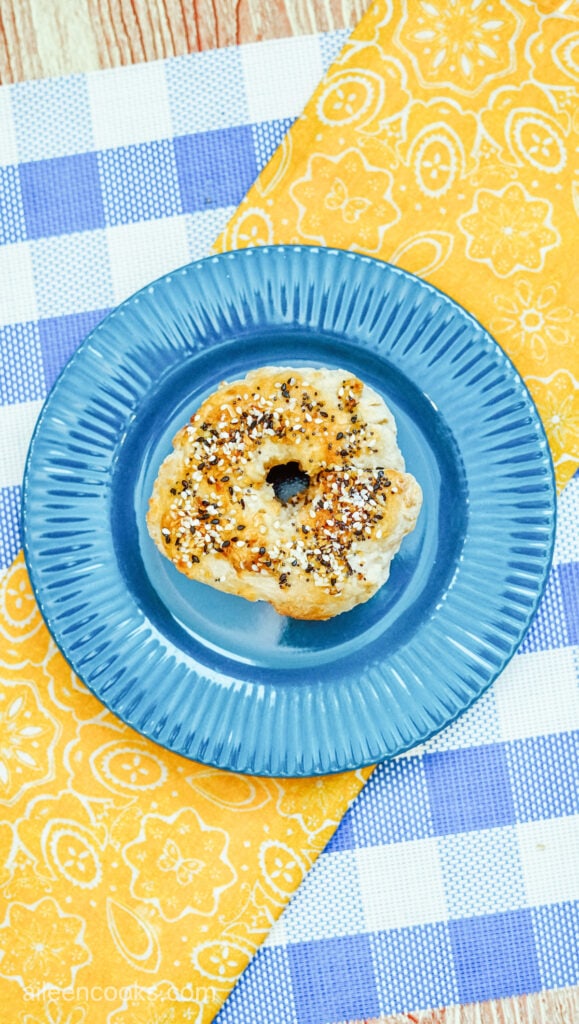 An everything bagel made in the air fryer, sitting on a blue plate with a yellow and blue tablecloth