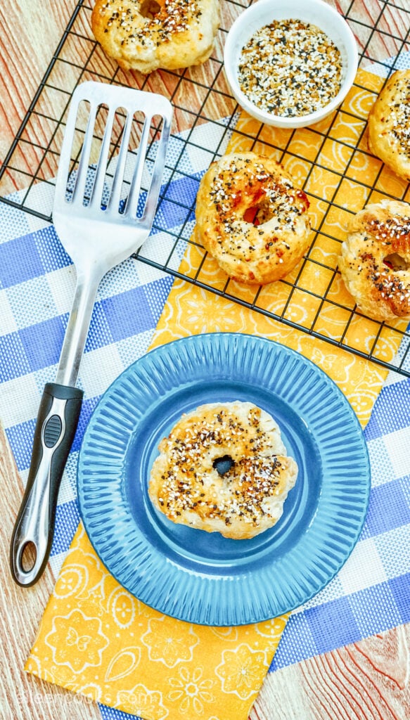 An air fryer bagel with everything bagel seasoning sitting on a blue plate with a spatula beside it, along with more bagels on a wire rack in the background
