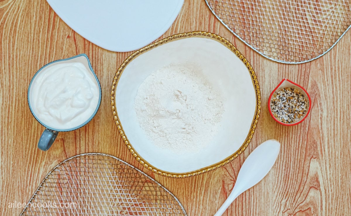 Flour in a white bowl, surrounded by additional ingredients like Greek yogurt and everything bagel seasoning