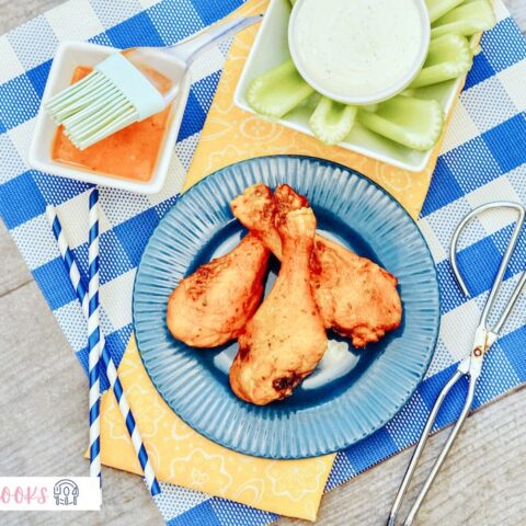 A serving of Air Fryer Buffalo Chicken on a blue round plate, sitting on a blue plaid place mat, surrounded by celery and small kitchen tools.