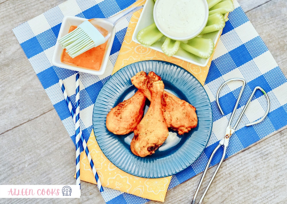 A serving of Air Fryer Buffalo Chicken on a blue round plate, sitting on a blue plaid place mat, surrounded by celery and small kitchen tools.