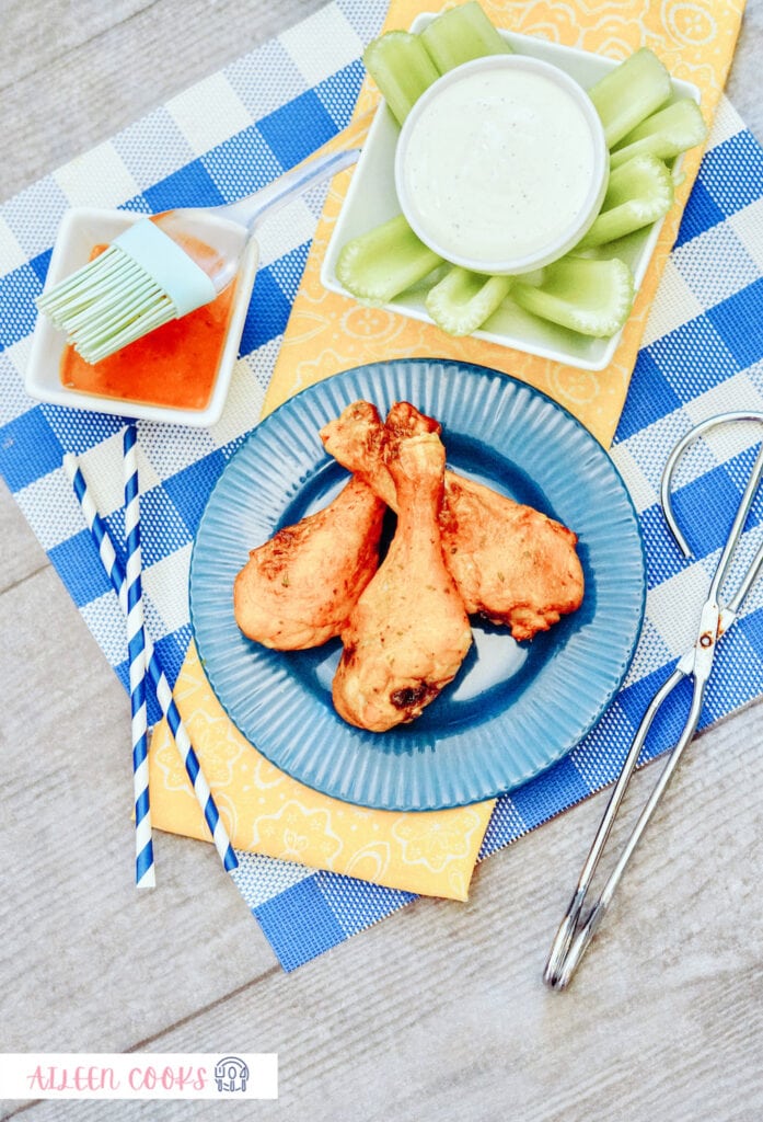 Bird’s eye view of Air Fryer Buffalo Chicken on a blue round plate, sitting on a blue plaid place mat, surrounded by celery and small kitchen tools.