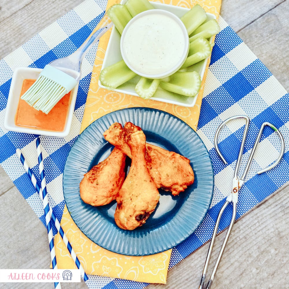 Bird’s eye view of Air Fryer Buffalo Chicken on a blue round plate, sitting on a blue plaid place mat, surrounded by celery and small kitchen tools.