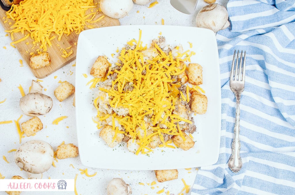 Ariel view of air fryer tater tot casserole served on a white plate with ingredients in the background, and a fork and a blue napkin on the side