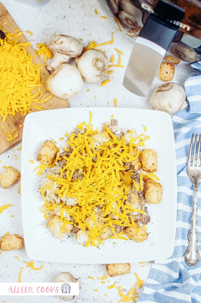 Ariel view of air fryer tater tot casserole served on a white plate with ingredients in the background, and a fork and a blue napkin on the side