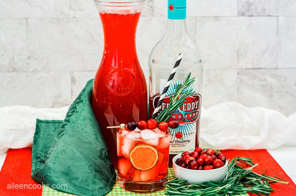 A cranberry Moscow mule, on red and green place mats with a marbled background, surrounded by additional Moscow mule ingredients
