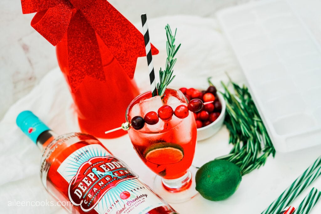 A Christmas Moscow Mule topped with cranberries and rosemary, on a white background, surrounded by vodka, an ice tray, and a lime.