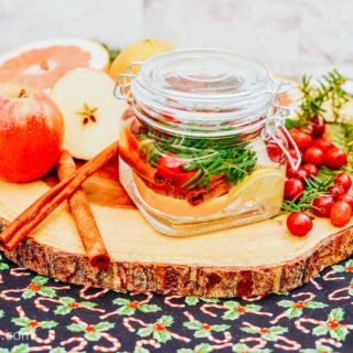 A holiday simmer pot poured into a small glass jar, sitting on top of a wooden board and on a holiday placemat, surrounded by ingredients like cranberries, apples, and cinnamon sticks