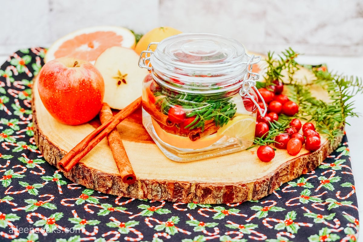 A holiday simmer pot poured into a small glass jar, sitting on top of a wooden board and on a holiday placemat, surrounded by ingredients like cranberries, apples, and cinnamon sticks