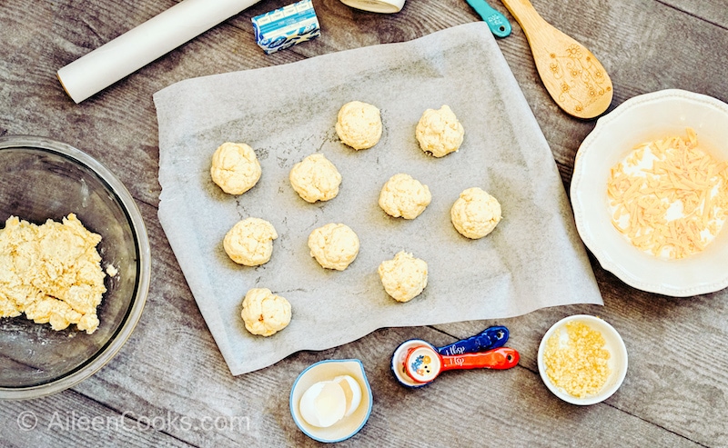 Unbaked Cheddar Bay Biscuits on a parchment-lined baking sheet, surrounded by small kitchen tools and a bowl of biscuit batter.
