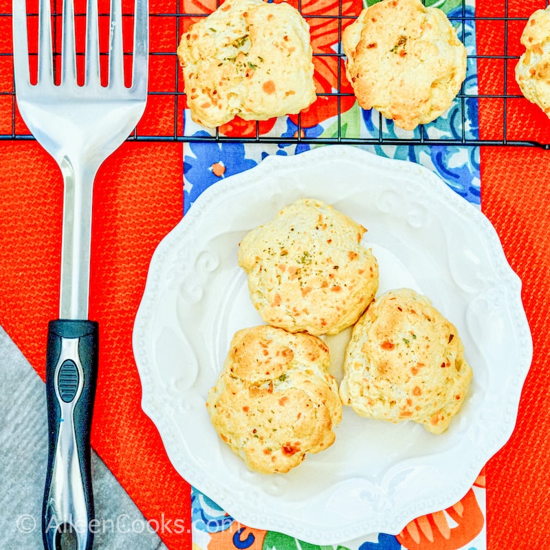 Cheddar Bay Biscuits in a white bowl, sitting on a red table cloth with additional biscuits in the background, sitting on a wire rack