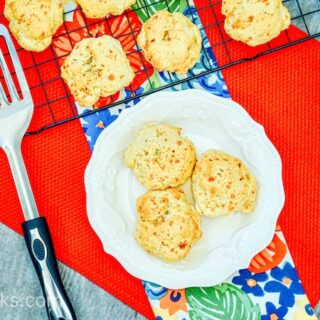Cheddar Bay Biscuits in a white bowl, sitting on a red table cloth with additional biscuits in the background, sitting on a wire rack